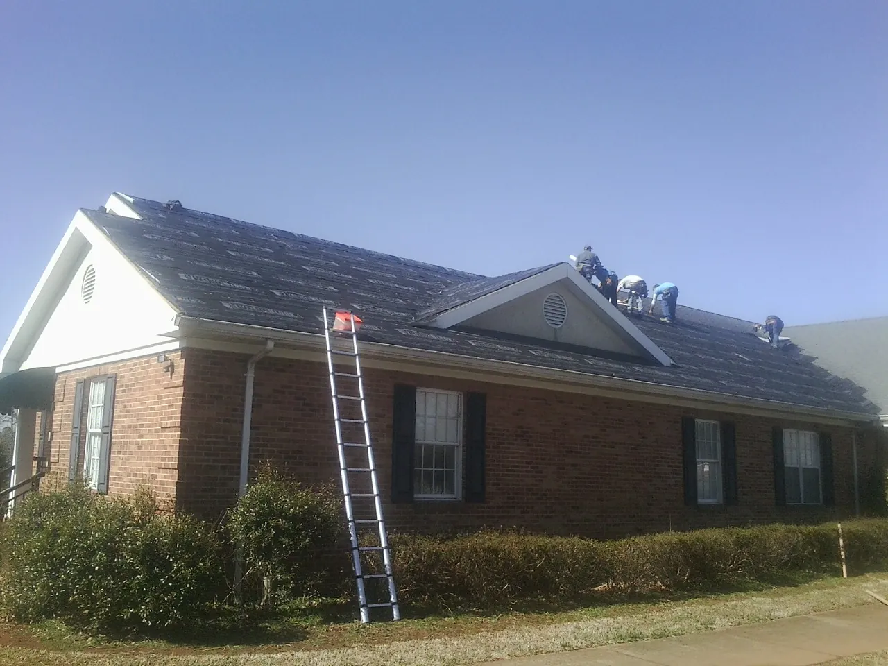 Crew tearing shingles off a brick ranch roof with a ladder set — Emergency Roof Repair work by our crew in Caret