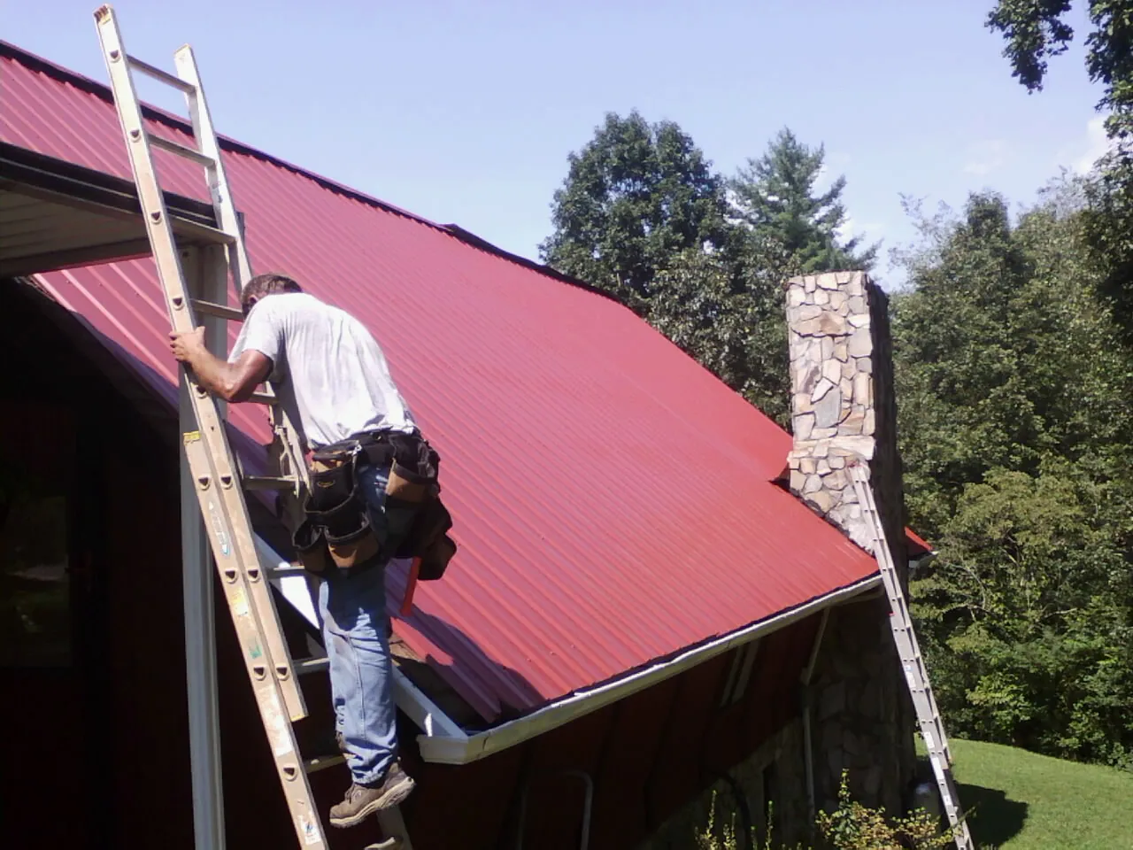 Roofer on a ladder beside a red metal roof and stone chimney — Soffit & Fascia Repair by Roofing Brothers in Caret